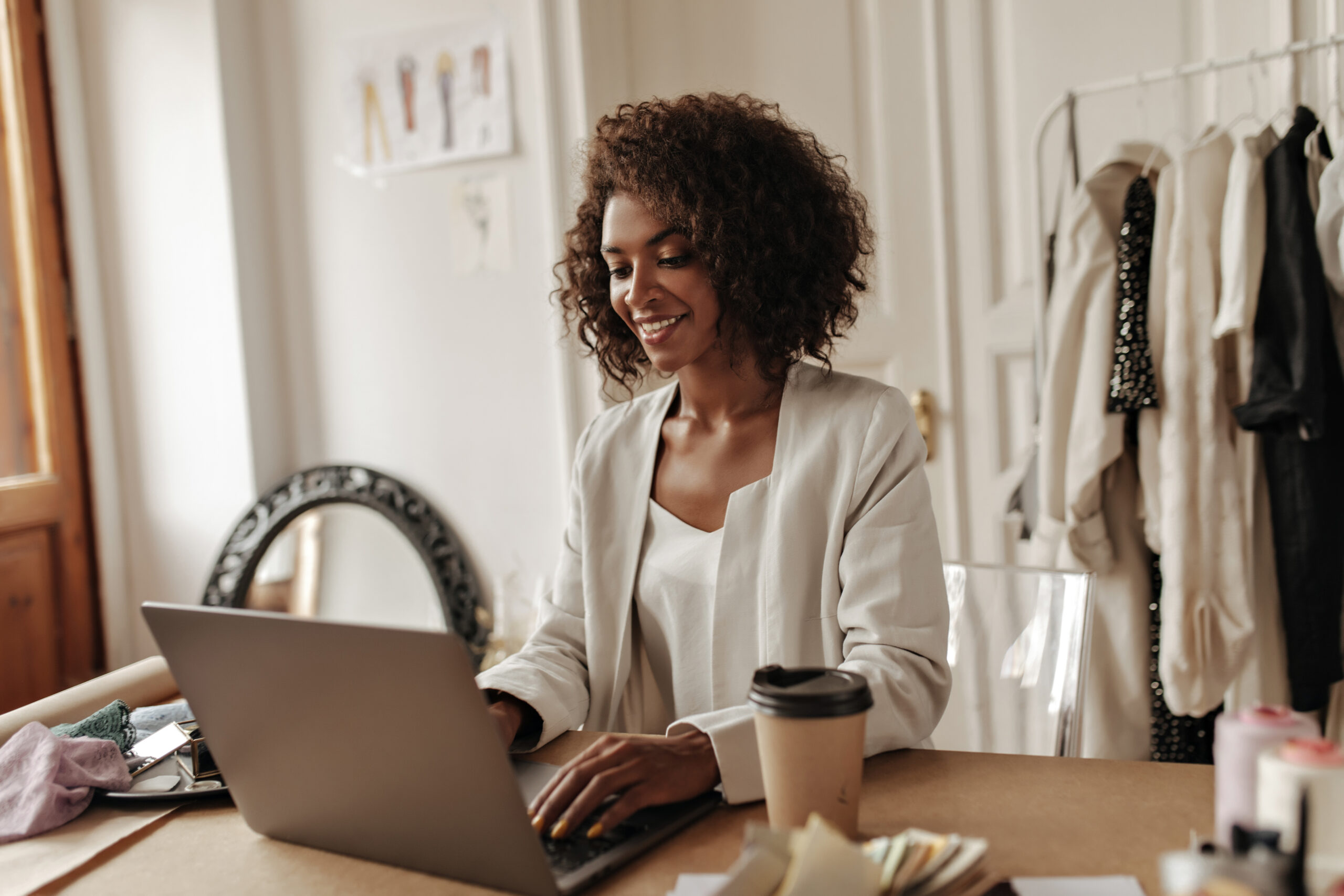 attractive curly darkskinned woman works laptop sits by desk with coffee cup happy designer poses cozy office